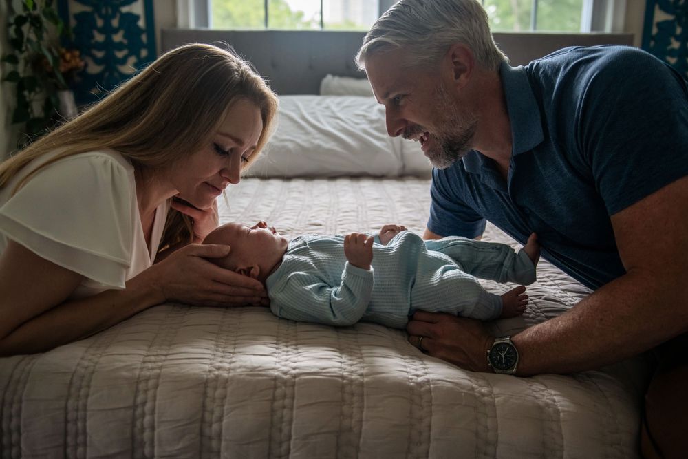 Parents lying on bed gazing adoringly at their newborn baby in soft natural light.