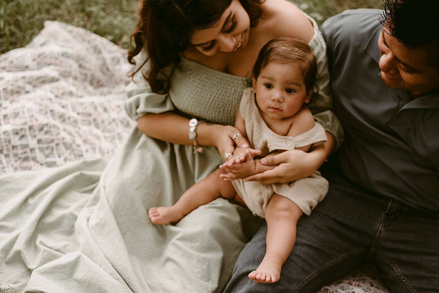 Tender family moment captured on lace blanket outdoors during golden hour photography session.