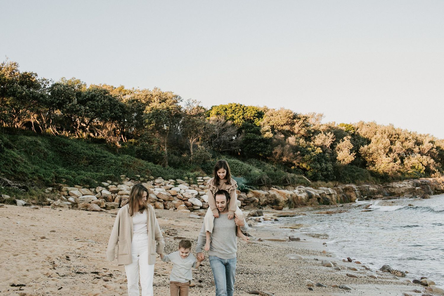 Family of four walks along rocky beach shoreline at golden hour with child on fathers shoulders.