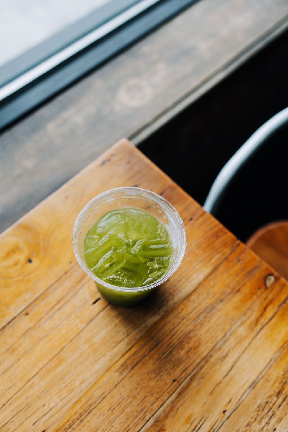 Green smoothie in a clear plastic cup on a wooden table by a window.