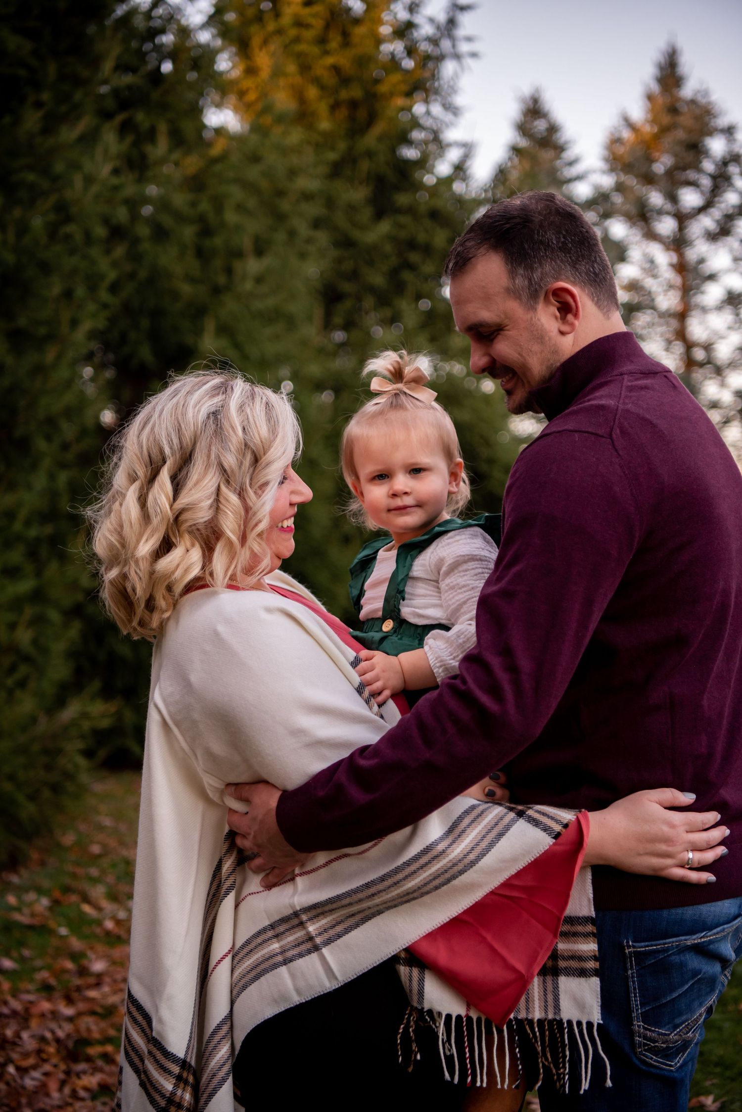 family of three with mom, dad and toddler girl in front of evergreen trees