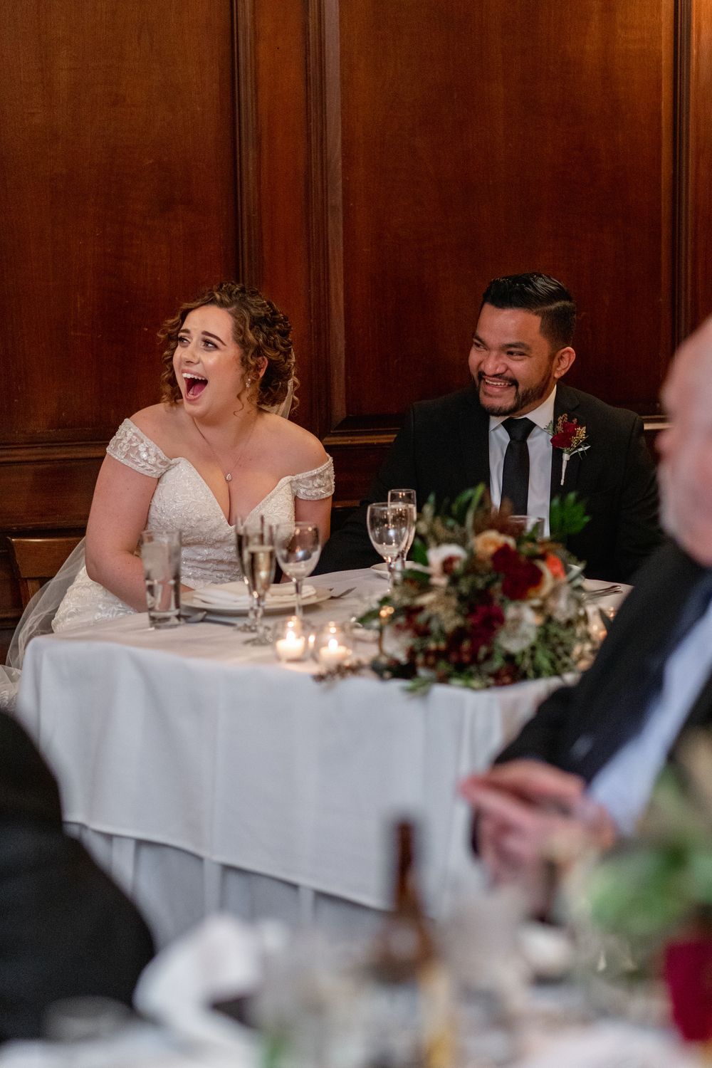 Wedding reception couple reacts joyfully to speeches at elegant dinner table.