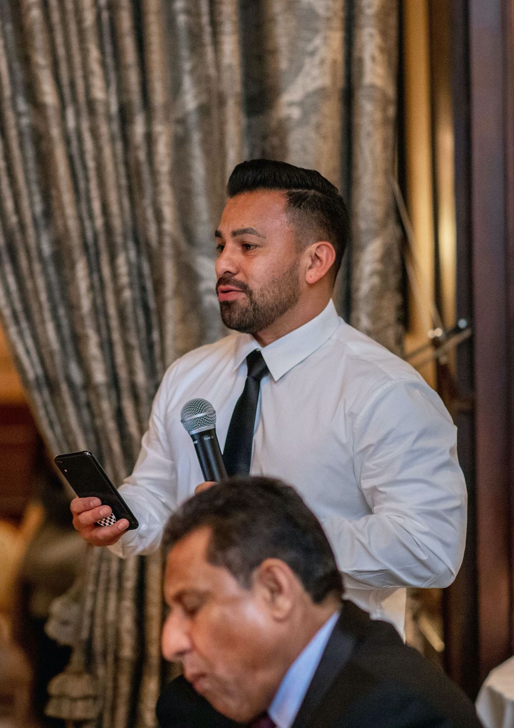 Wedding guest delivers speech while holding phone and microphone at reception dinner.