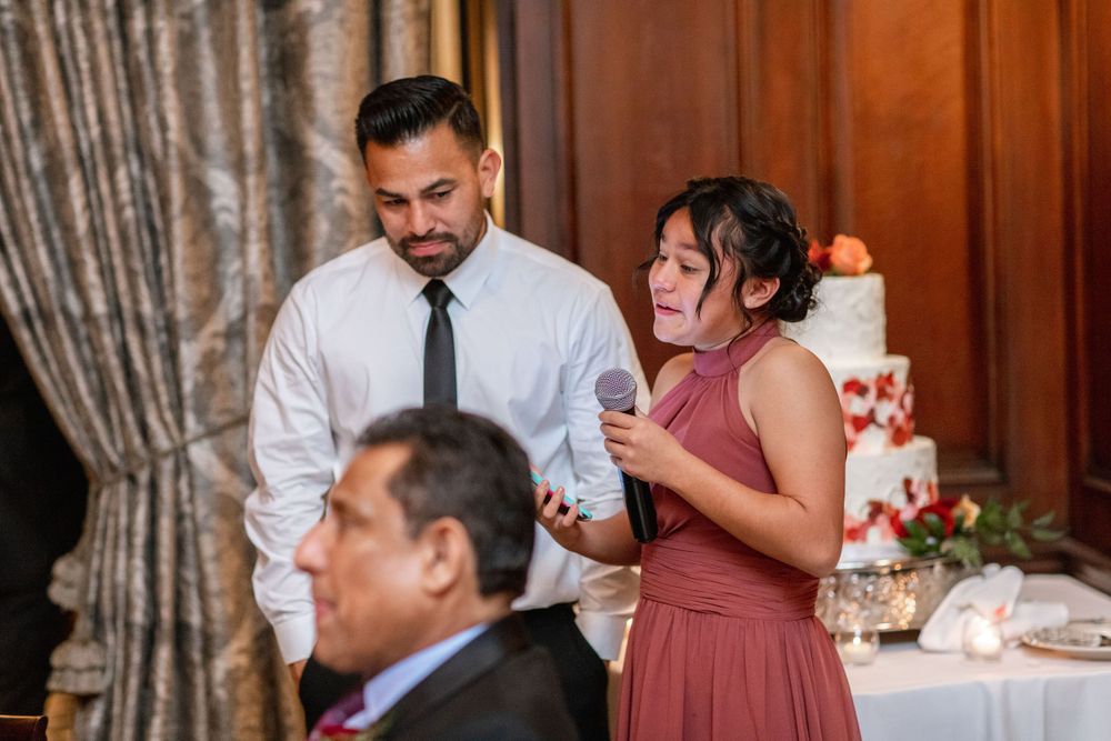Reception guests enjoy speeches and laughter during wedding dinner in elegant wood-paneled room.