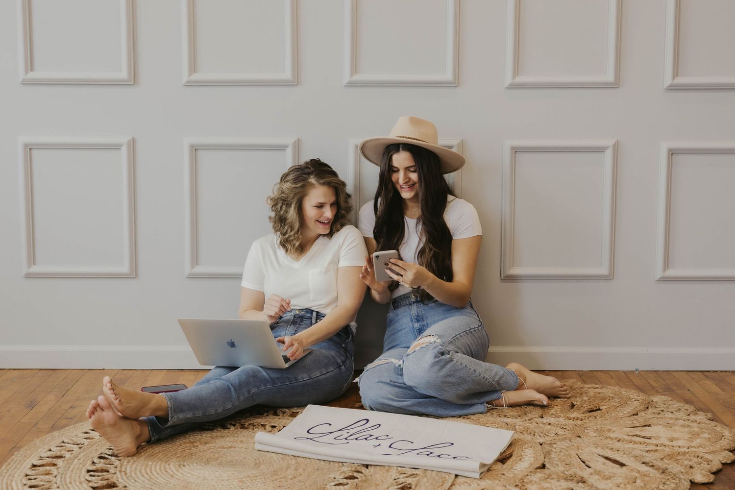 Two friends sit on a round rug looking at phones while wearing casual denim and a sun hat.