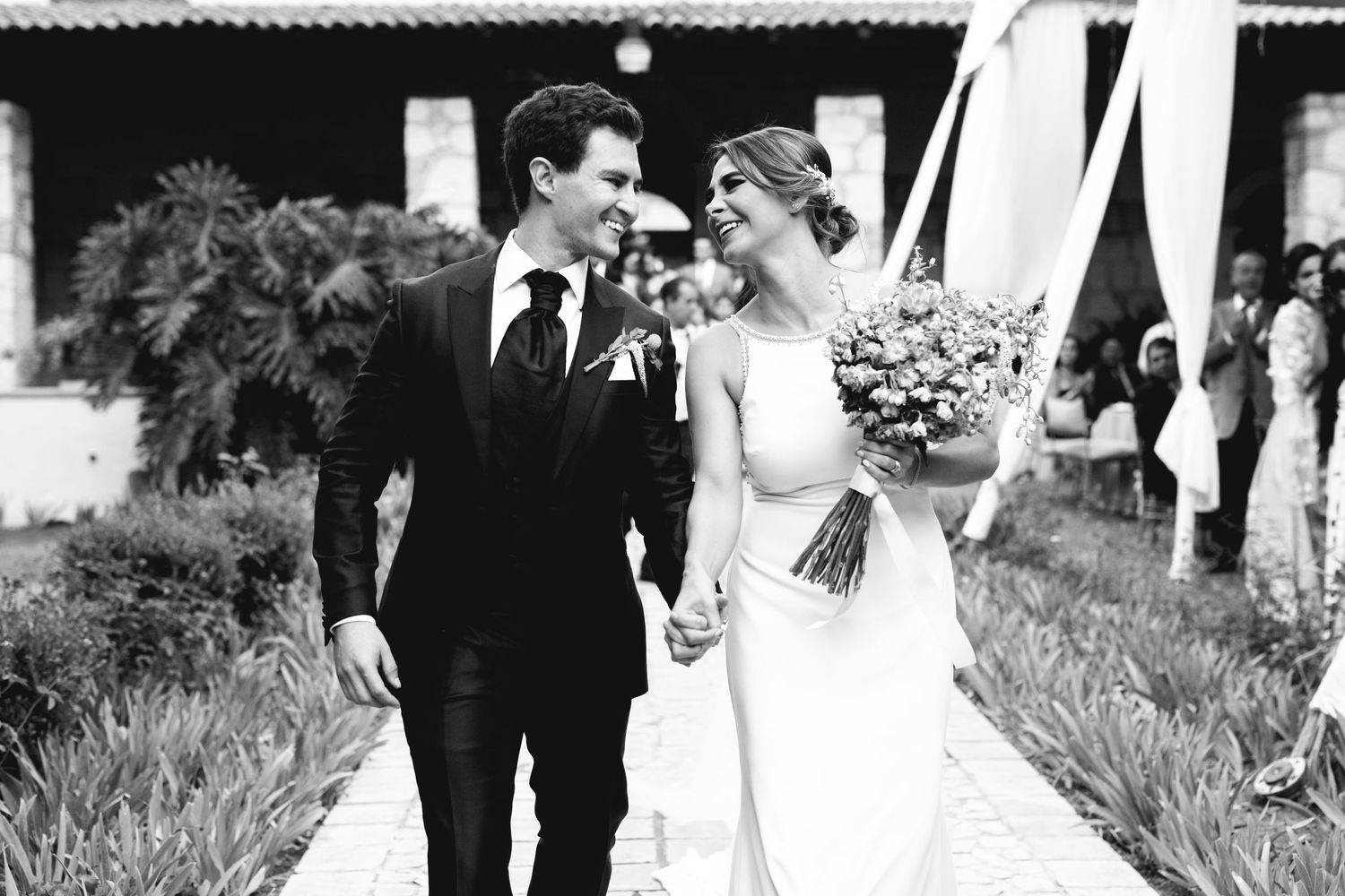 Black and white wedding photo of newlyweds walking together outdoors with floral bouquet.