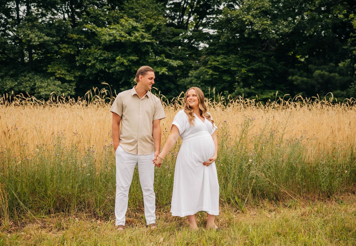 A couple looks at each other in a field of tall grass. The woman is pregnant and holds her belly. Captured by Grace