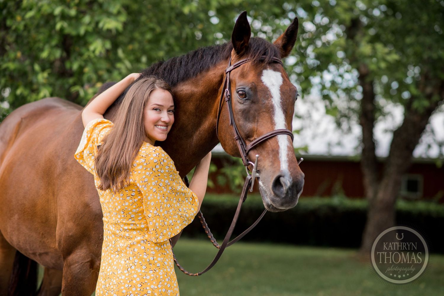A bay horse with white blaze stands next to person wearing yellow floral dress in garden setting.