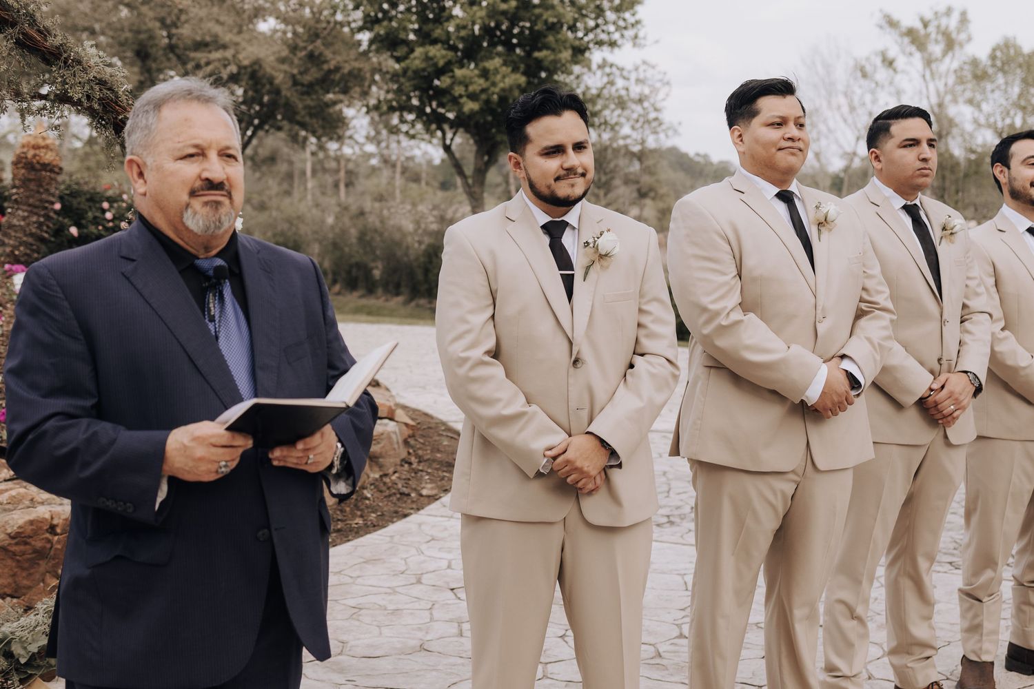 Wedding party with officiant in navy suit and groomsmen in beige suits stand during outdoor ceremony.
