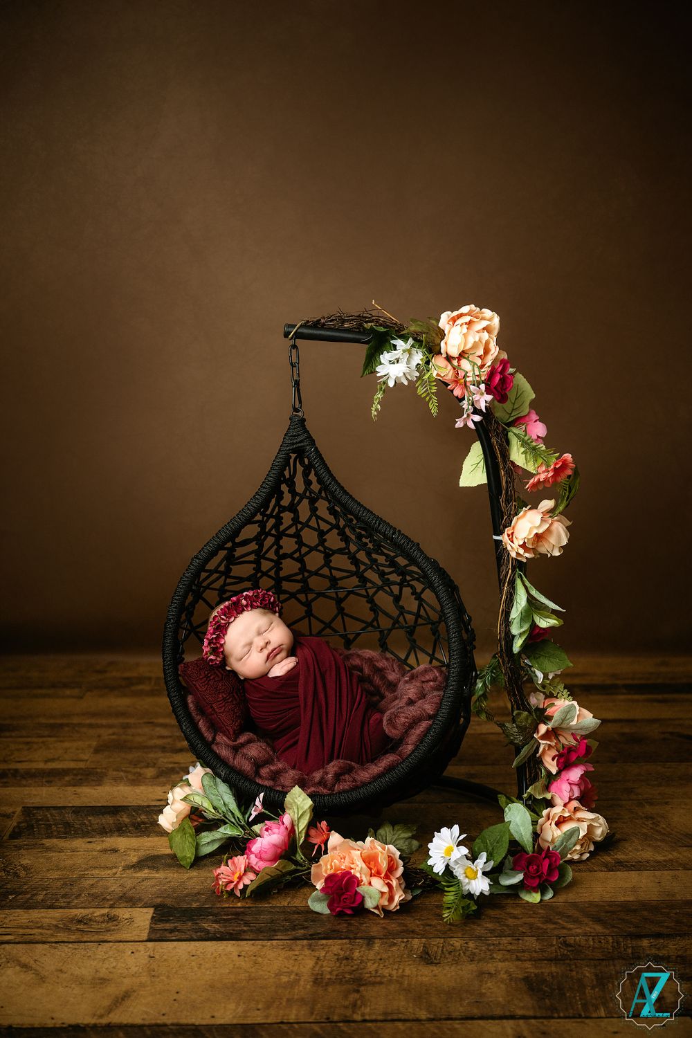 A hanging wicker pod chair decorated with floral garlands cradles a newborn wrapped in burgundy fabric.