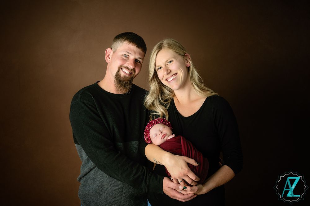 Professional studio portrait of parents in black holding their newborn against a dark brown backdrop.