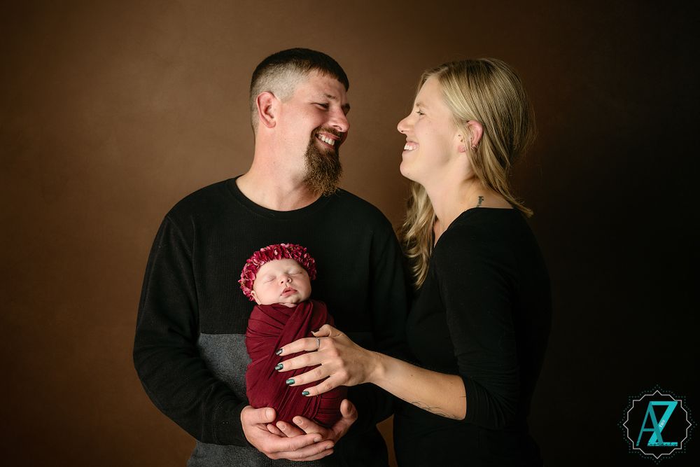 Studio photograph shows parents sharing a joyful moment while holding their newborn in burgundy wrap.
