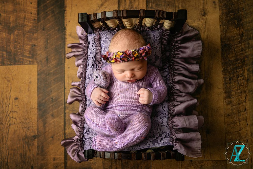 Sleeping newborn in purple wrap lies on ruffled cushion wearing delicate floral crown.