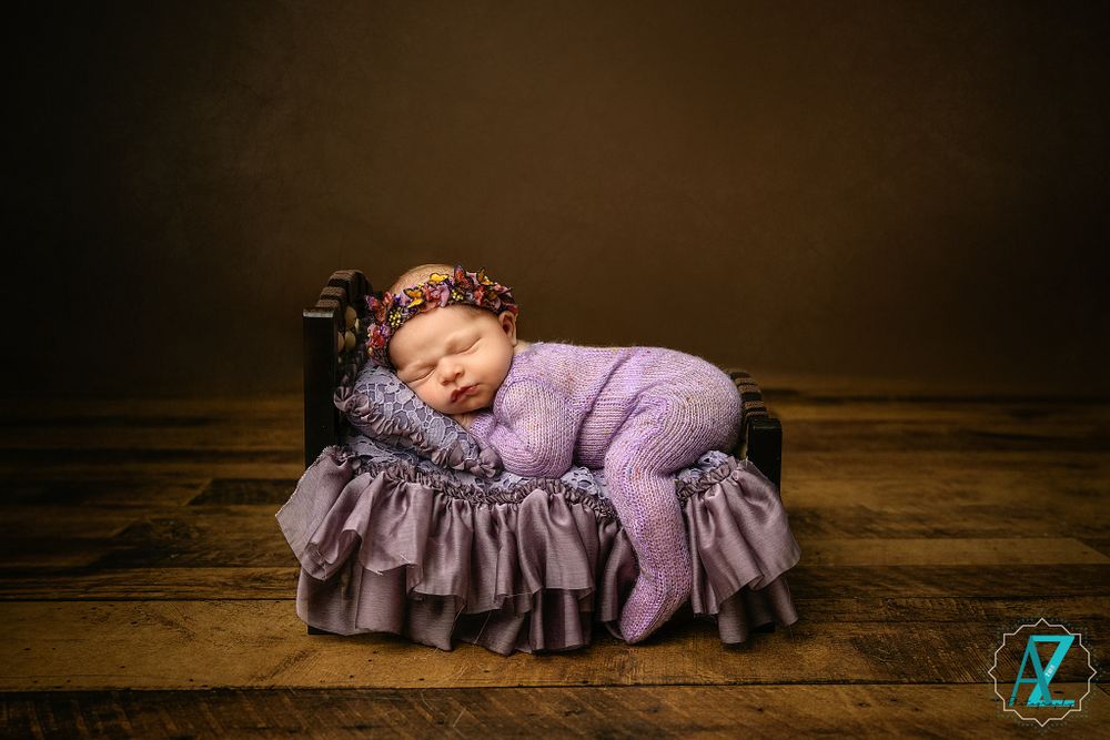 Newborn wearing purple ruffled outfit rests peacefully on matching miniature chaise lounge prop.