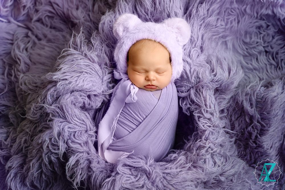 Sleeping newborn wrapped in purple fabric wearing bear ear hat rests on fluffy violet backdrop.
