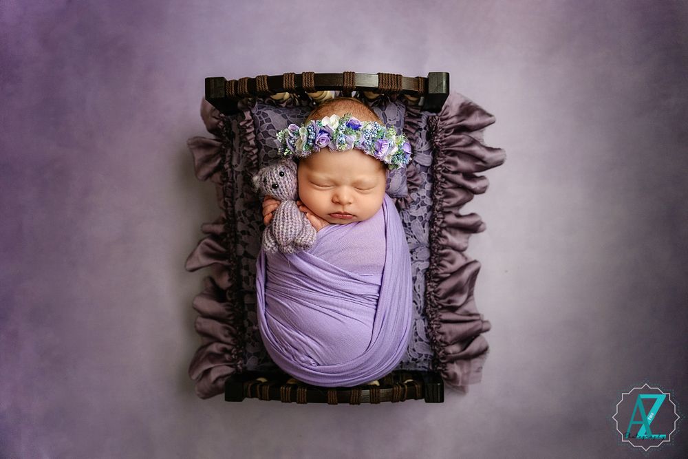 Overhead view of sleeping newborn wrapped in purple on dark ruffled cushion with floral headpiece.
