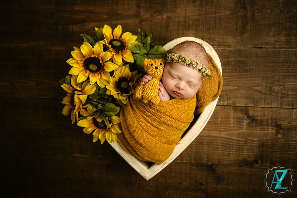 Newborn wrapped in yellow fabric rests in heart-shaped basket with sunflowers on wooden background.