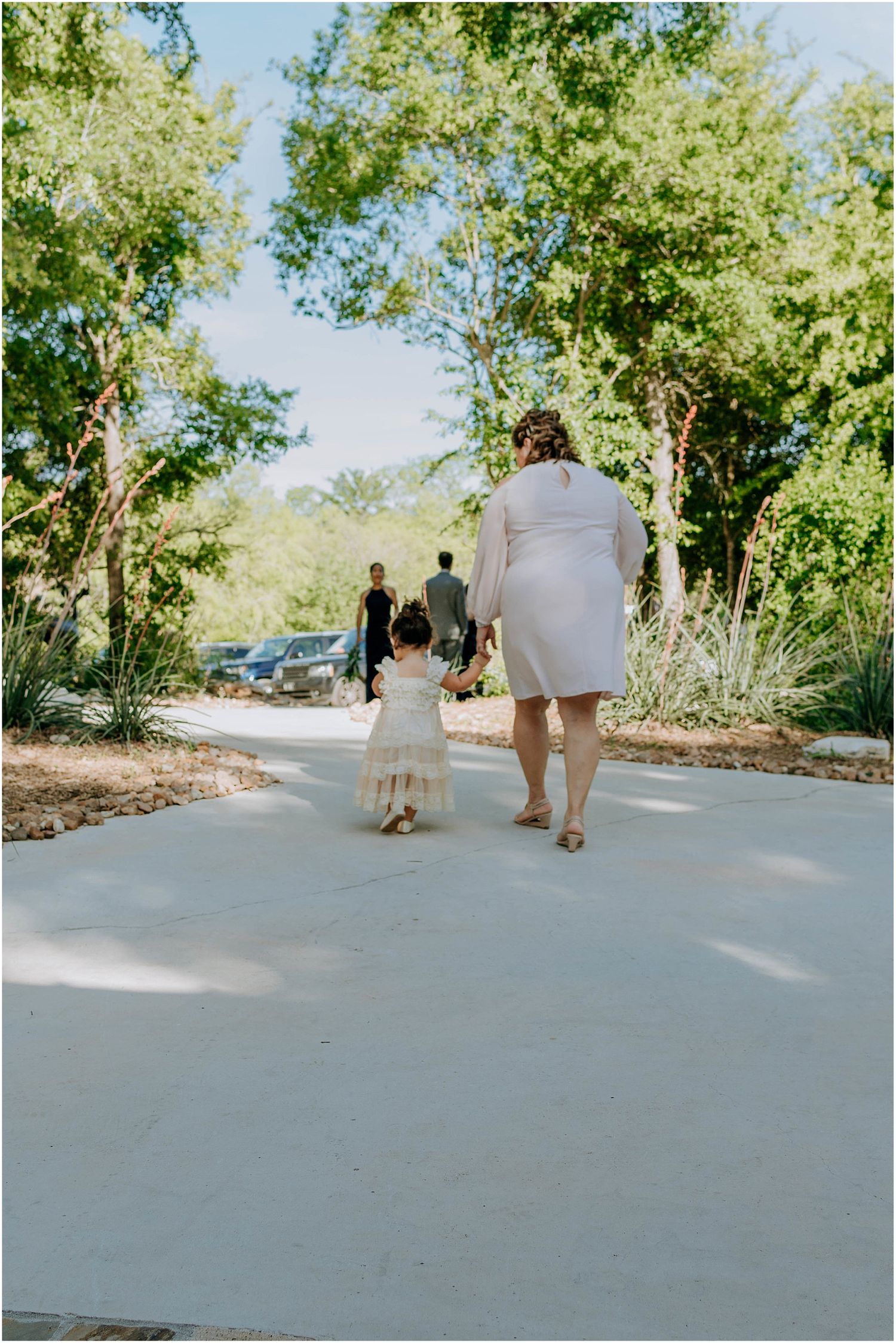 A sweet moment captured as two people walk along a sunny concrete path surrounded by greenery.