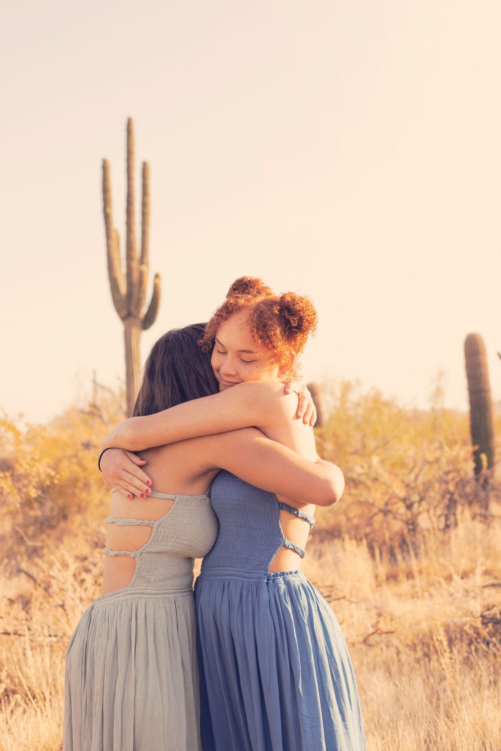Two people in flowing dresses embrace warmly in a desert setting with saguaro cacti.