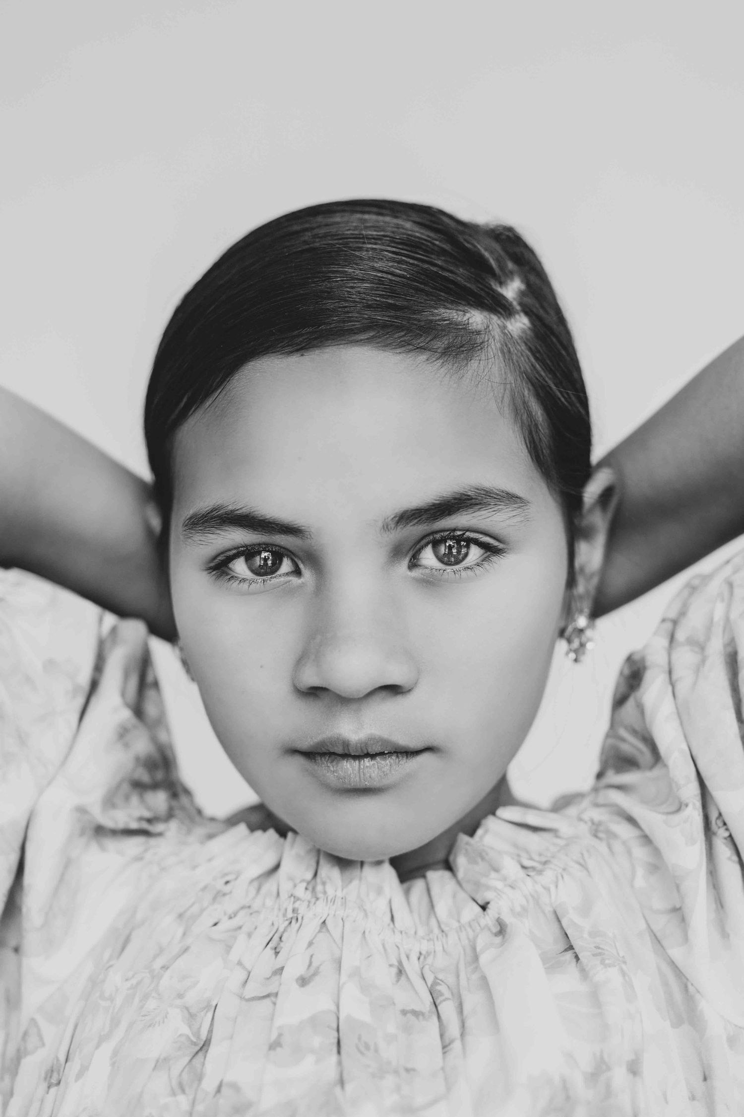 A black and white portrait of a young girl with slicked-back hair posing with hands behind her head.