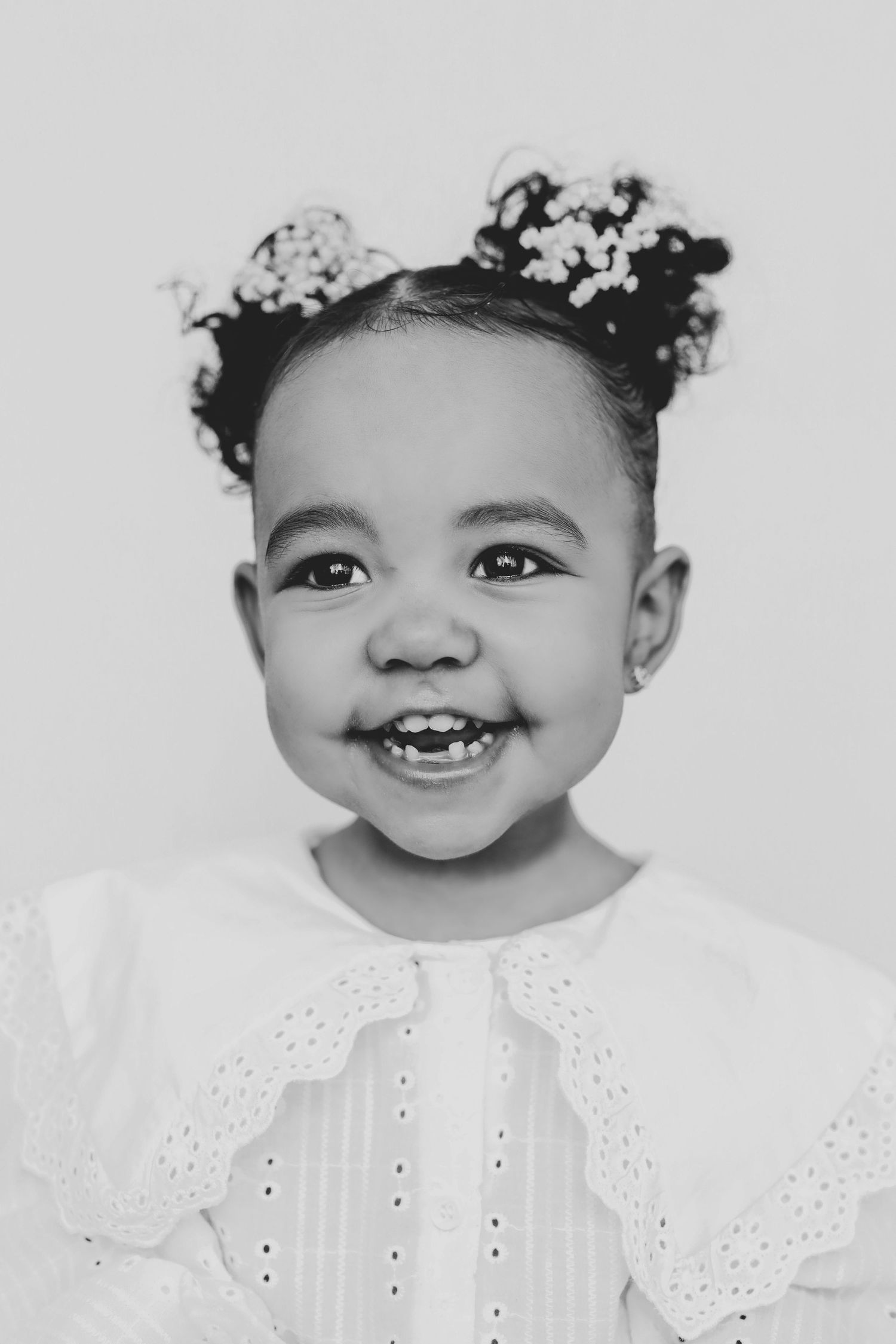 A black and white portrait of a smiling toddler girl with floral hair buns in a white dress.