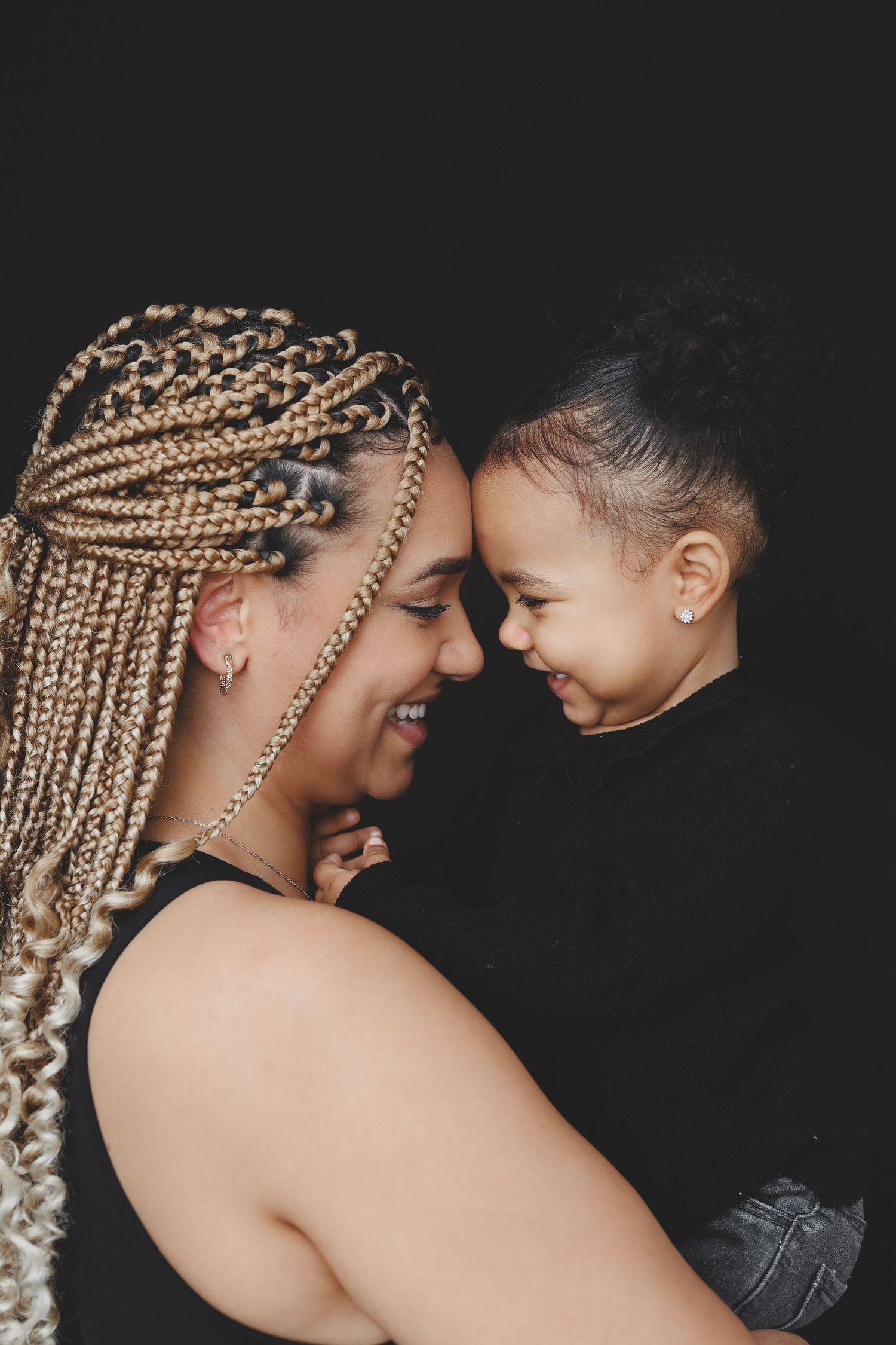 A joyful mother with blonde braids touches foreheads with her smiling toddler against a dark background.