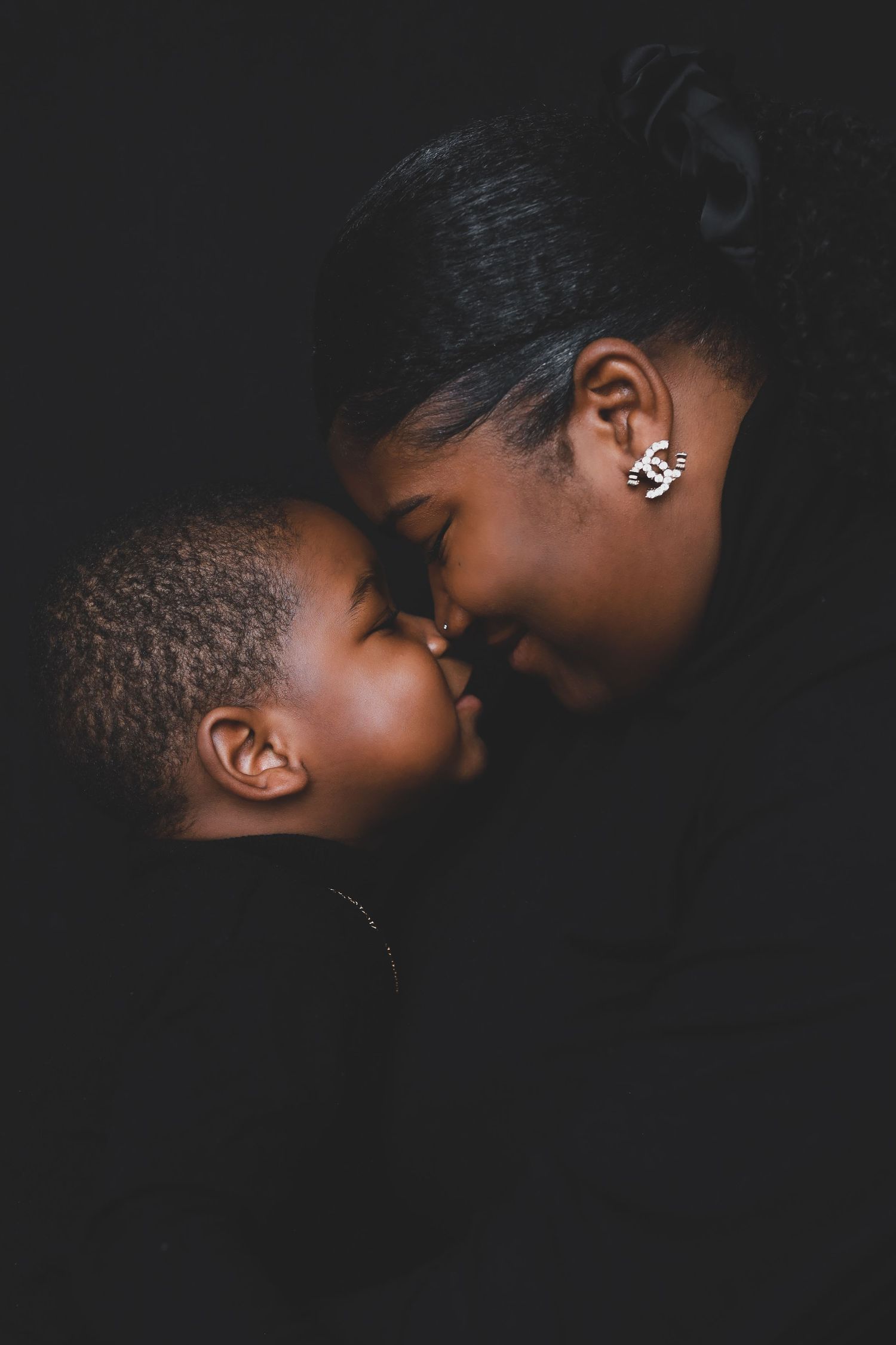 A tender moment between a mother and young child touching foreheads in an intimate dark studio portrait.