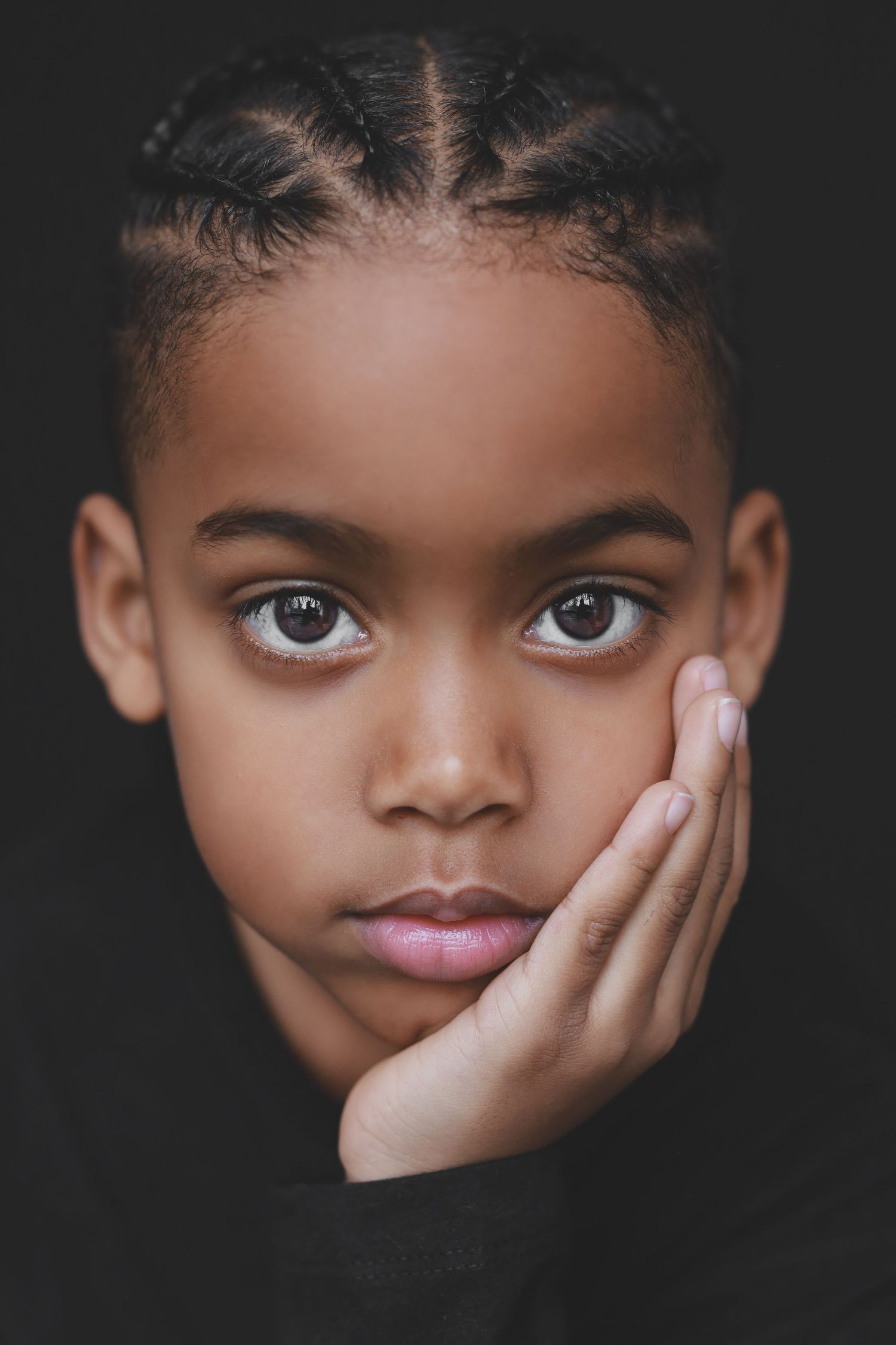 Portrait of a young Black girl with braided hair, resting hand on cheek, against a dark background.