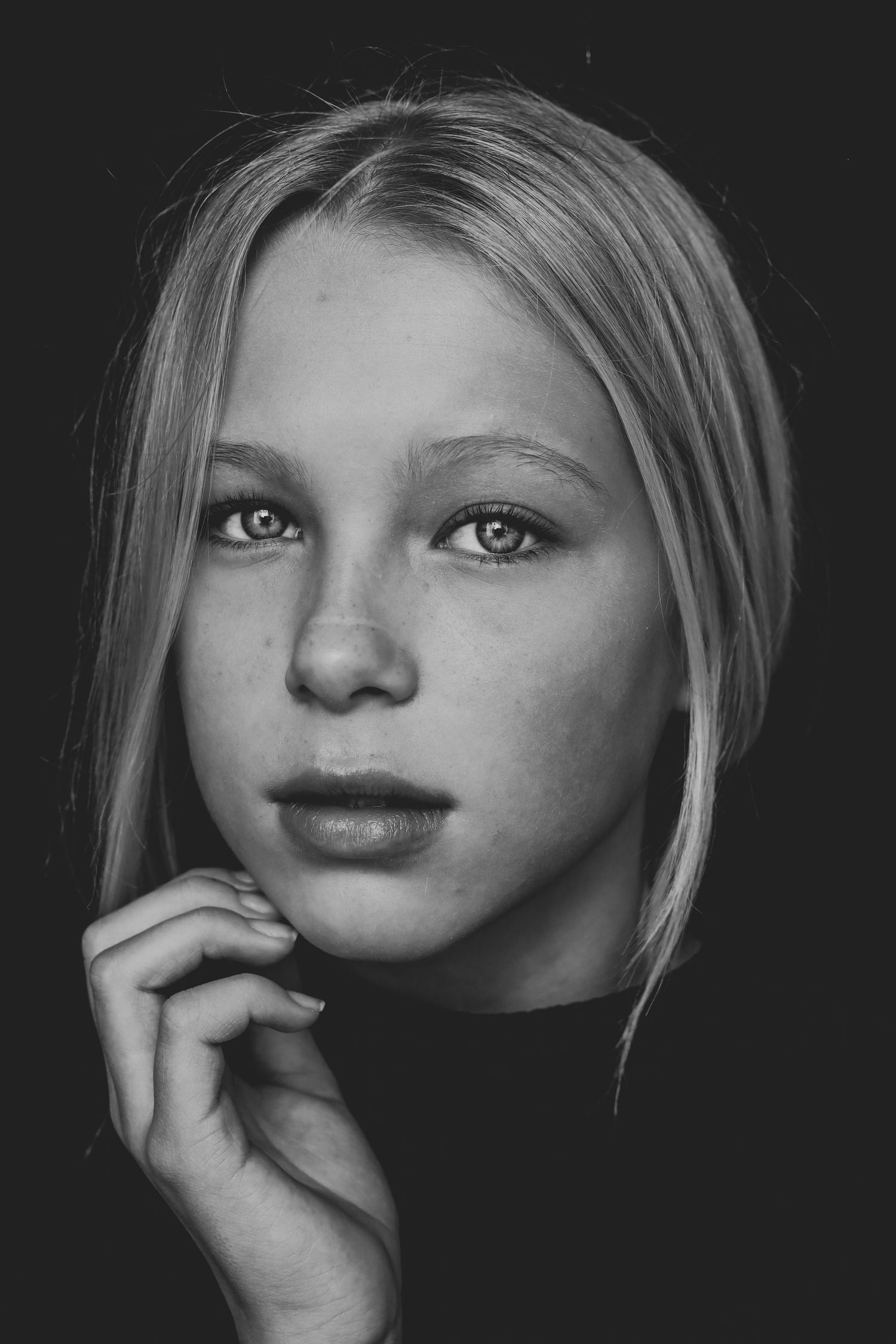 Black and white portrait of a blonde teenage girl with striking eyes, resting hand near chin on dark background.