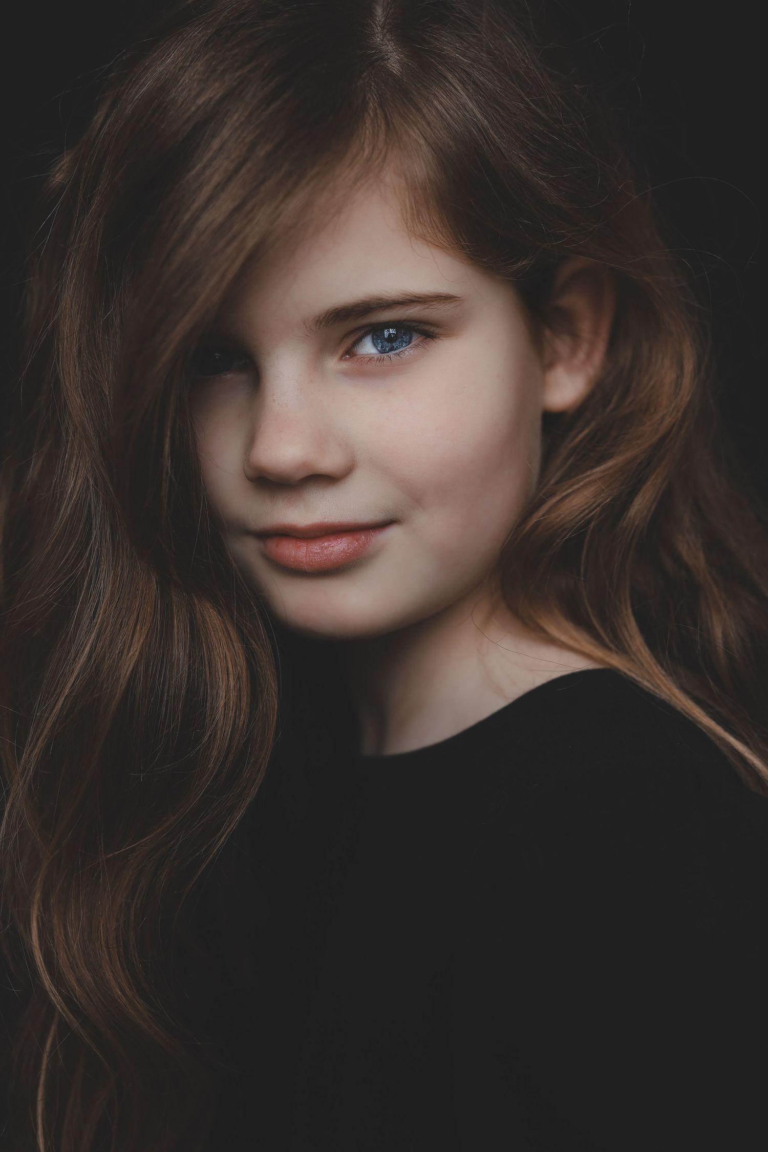A young girl with long brown hair and blue eyes smiles softly in a dark studio portrait.