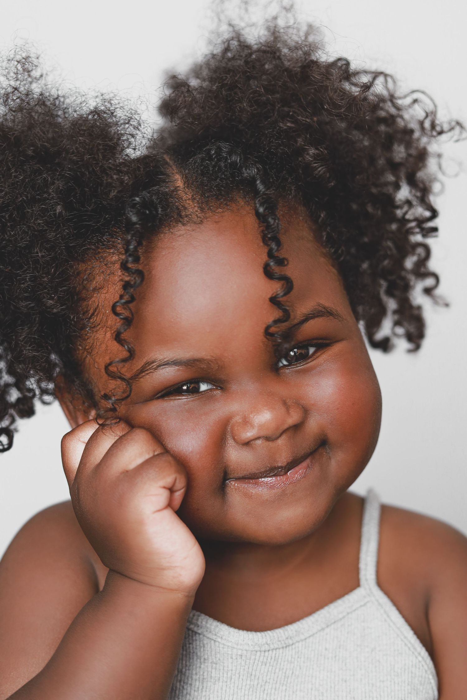 An adorable Black toddler girl with curly pigtails smiles playfully with her hand on her cheek.