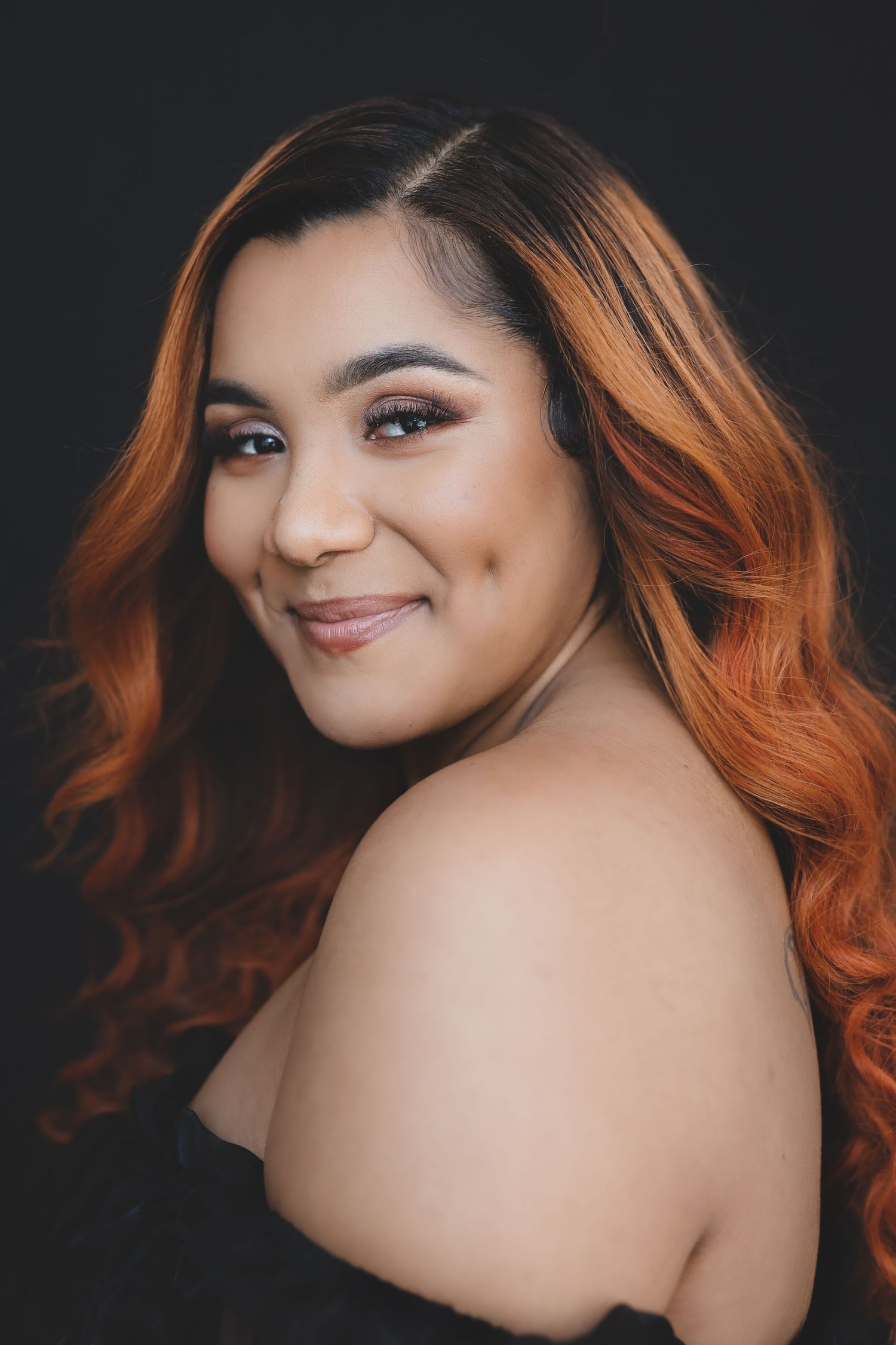 A plus-size woman with auburn ombre hair smiles over her shoulder in a studio portrait.