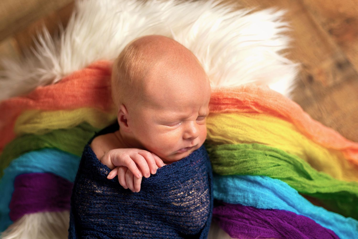 Sleeping newborn wrapped in navy blue fabric on a rainbow colored blanket.