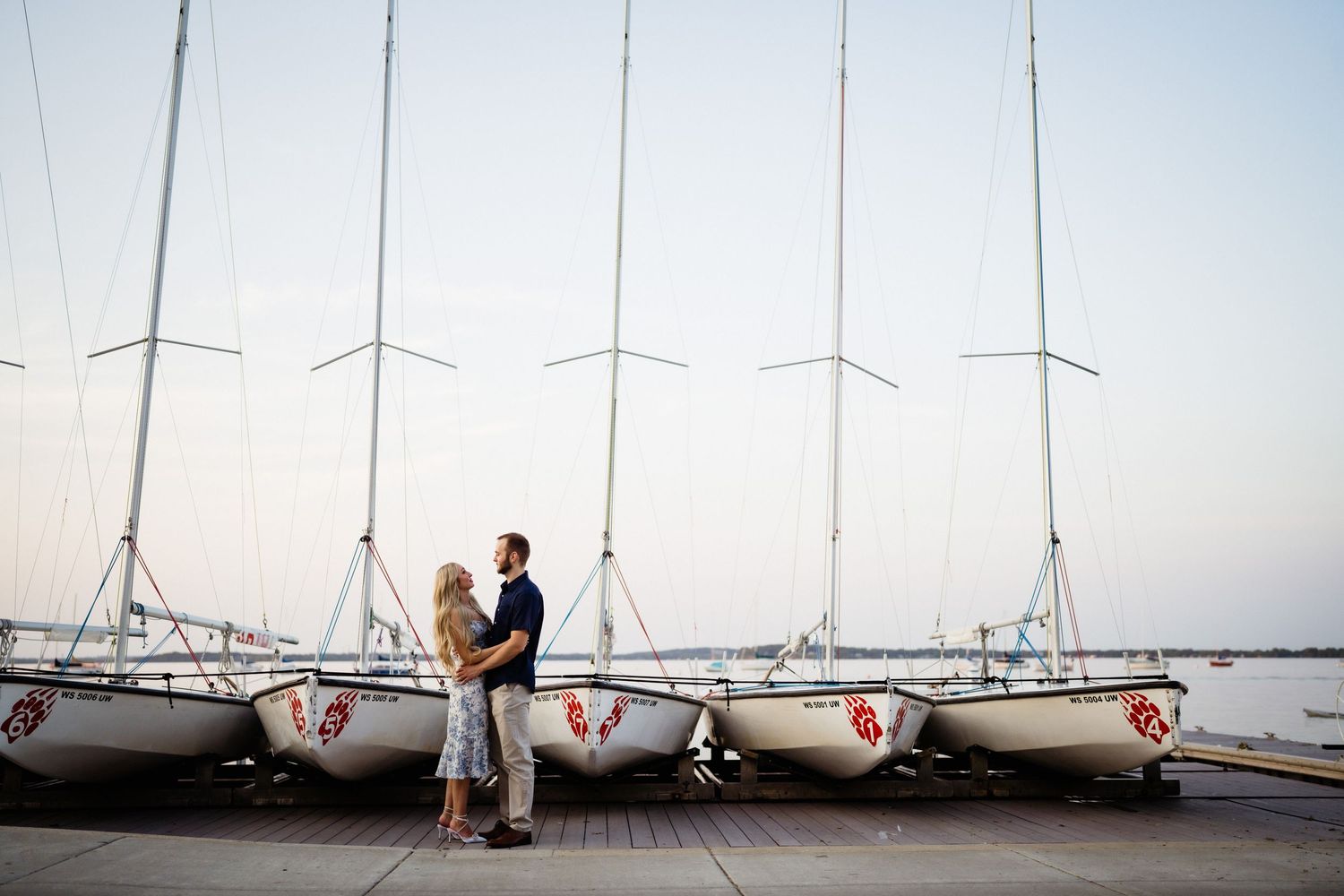 A couple stands close together on a dock with sailboats lined up at sunset.