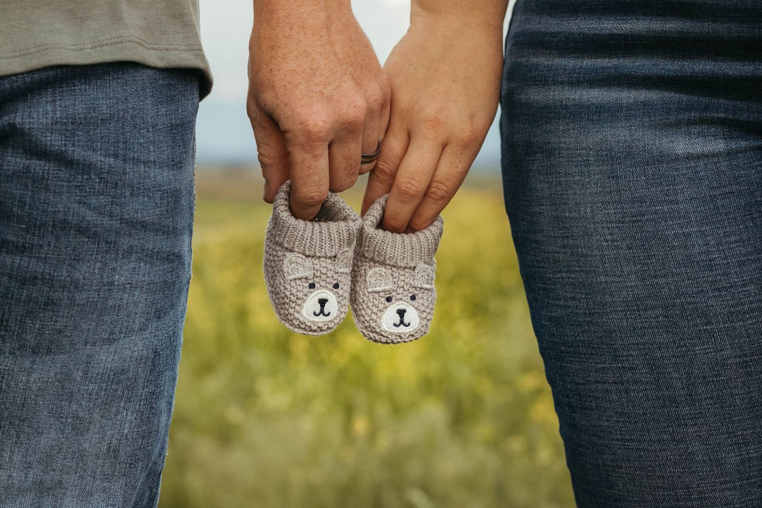 Hands holding tiny knitted bear baby booties against a blurred outdoor background.