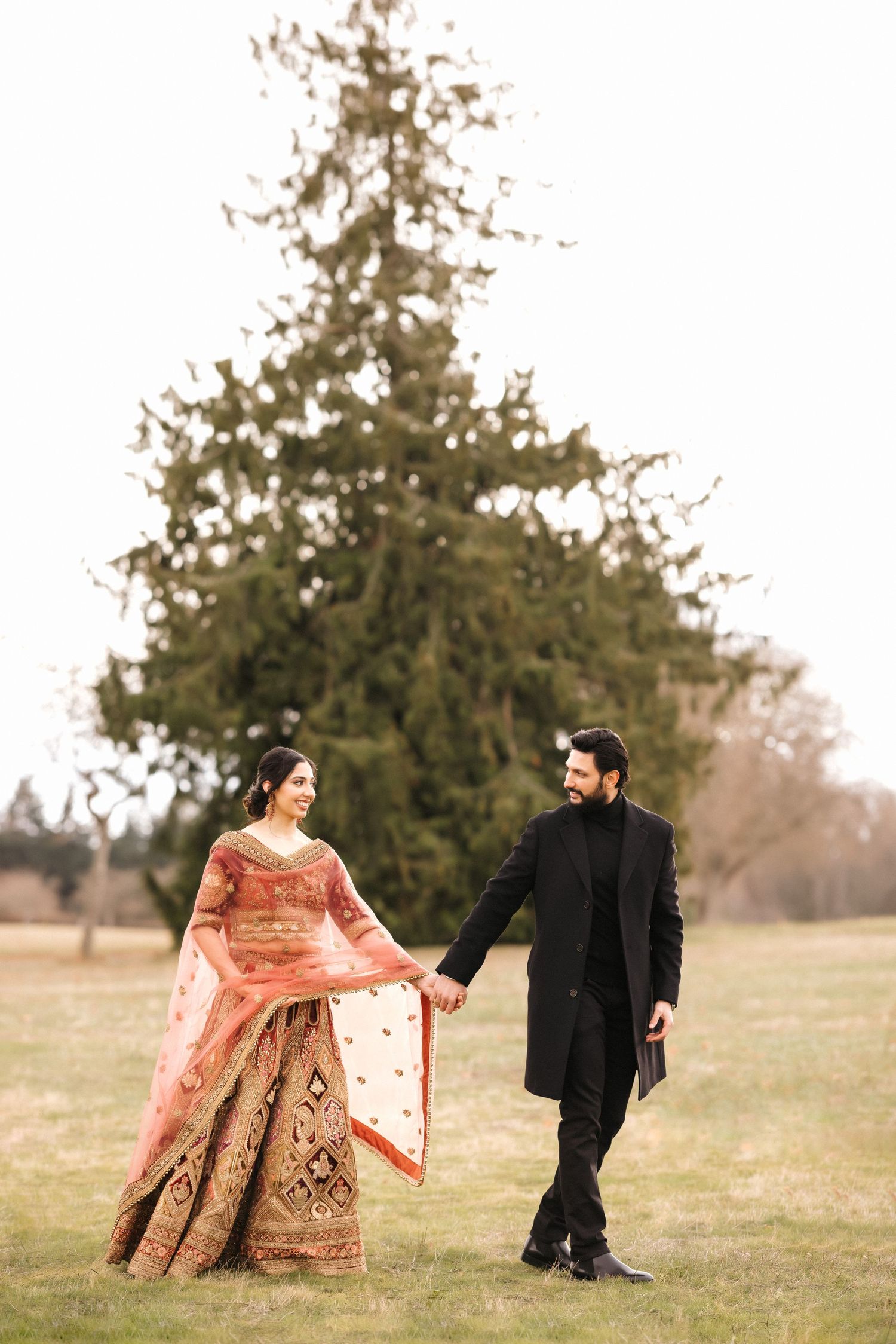 A couple in formal attire poses together in a field with evergreen trees in the background.