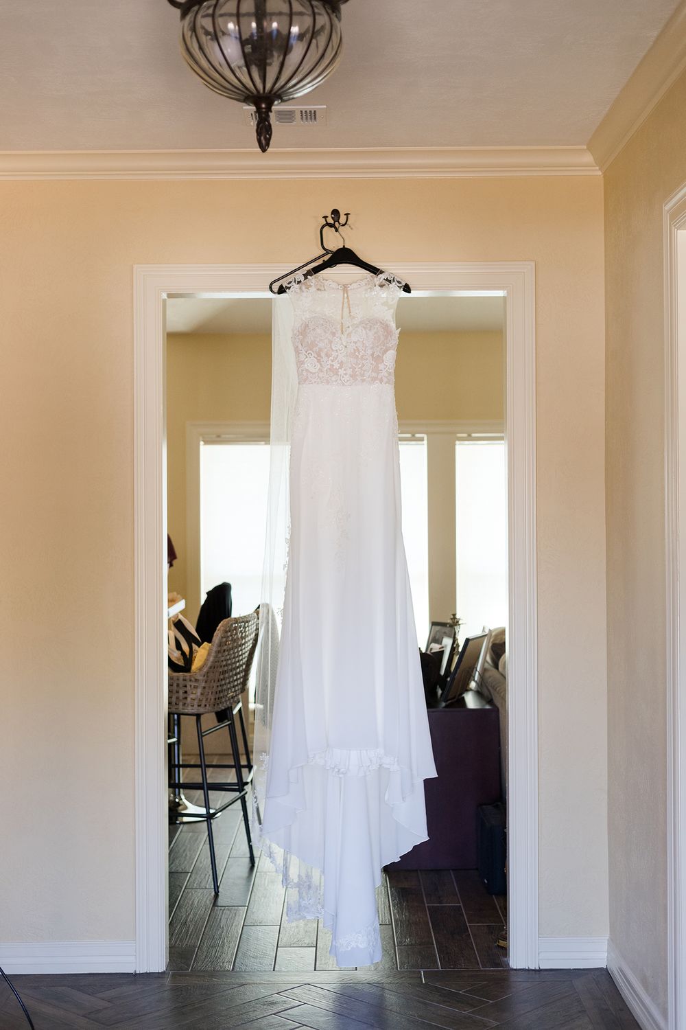 White lace wedding dress hanging in doorway with elegant light fixture above.