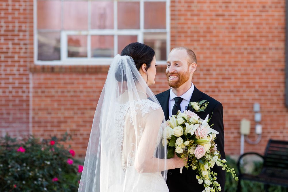 Romantic wedding portrait with bride holding white bouquet against brick backdrop.