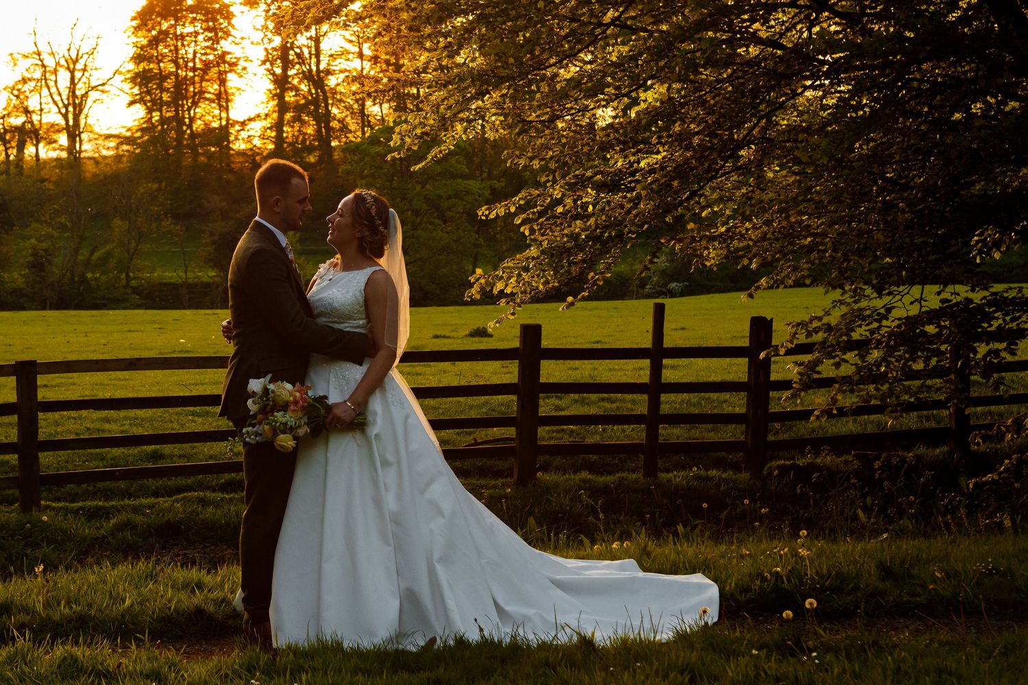 Golden sunset light illuminates couple by wooden fence in romantic countryside setting.