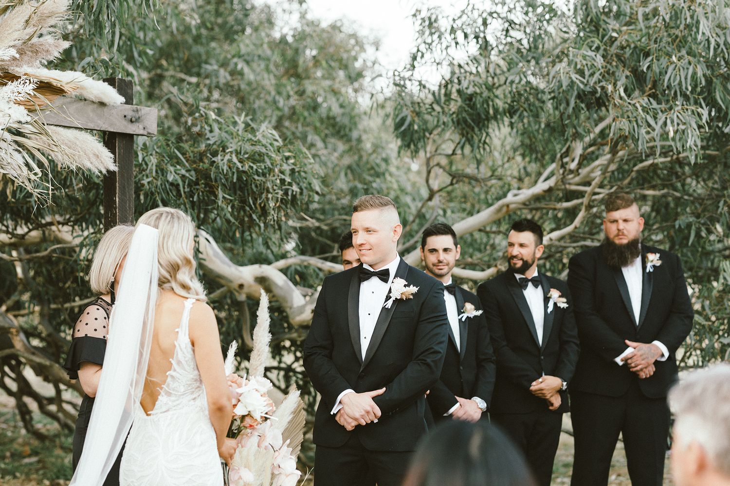 Wedding ceremony with groomsmen in black tuxedos under willow trees.