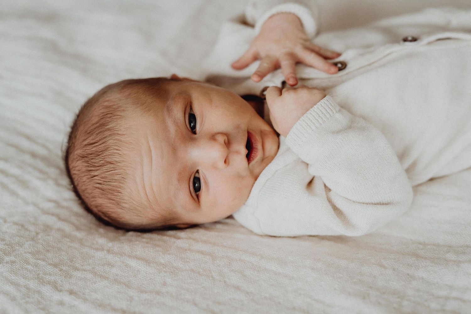 A baby in white clothing lying on soft white bedding looking at the camera.