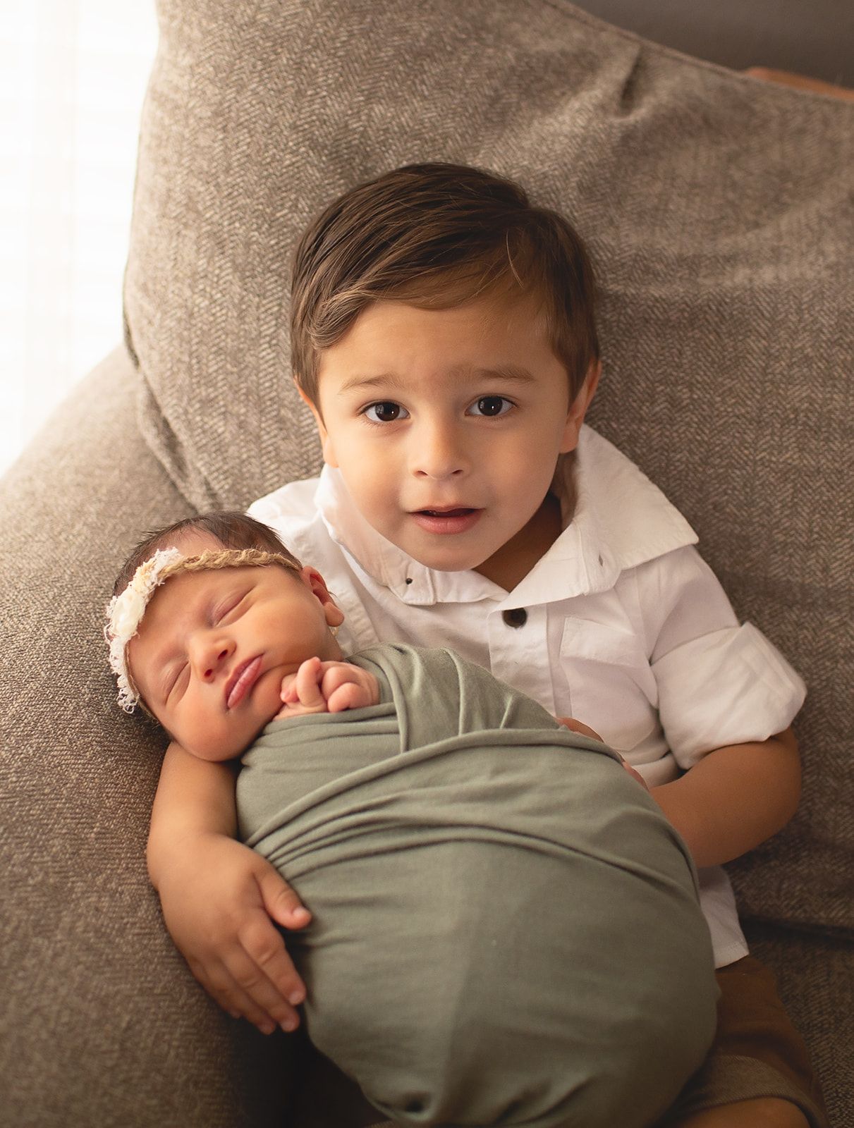 Two siblings share a tender moment on a gray couch as the older one in white hugs the swaddled baby in sage green.