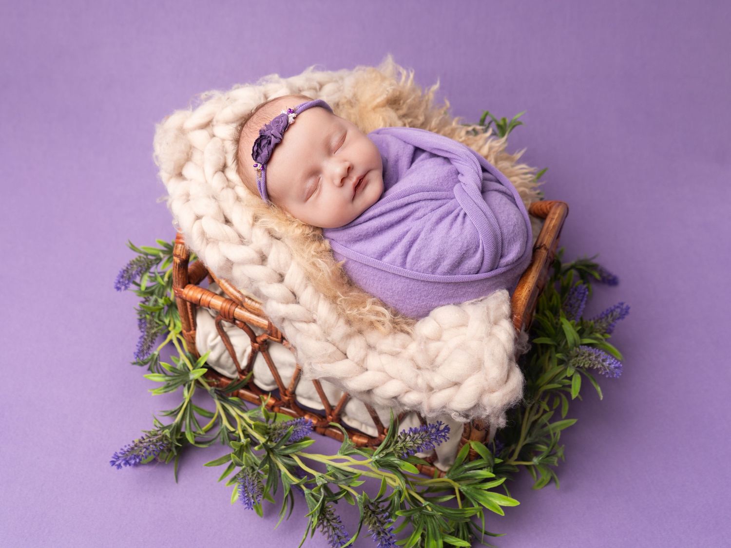 sleeping newborn baby girl in basket, fluffy blankets on purple backdrop with wrap client of Heather Katie Photography.