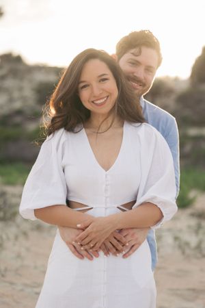 Joyful couple embracing and laughing together on sandy beach during sunset photoshoot.