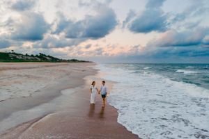 A couple walking along a beach shoreline during sunset with dramatic clouds overhead.