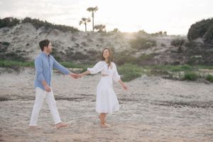 Couple in white outfits playing and dancing on sandy beach at sunset.