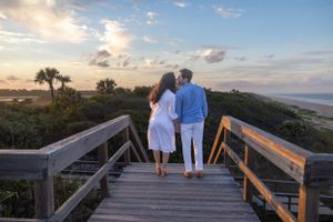 Couple standing on wooden boardwalk overlooking beach at sunset.