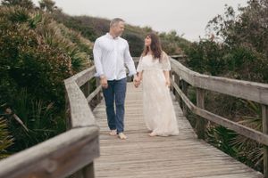 Couple walking hand in hand on a wooden boardwalk surrounded by coastal vegetation.