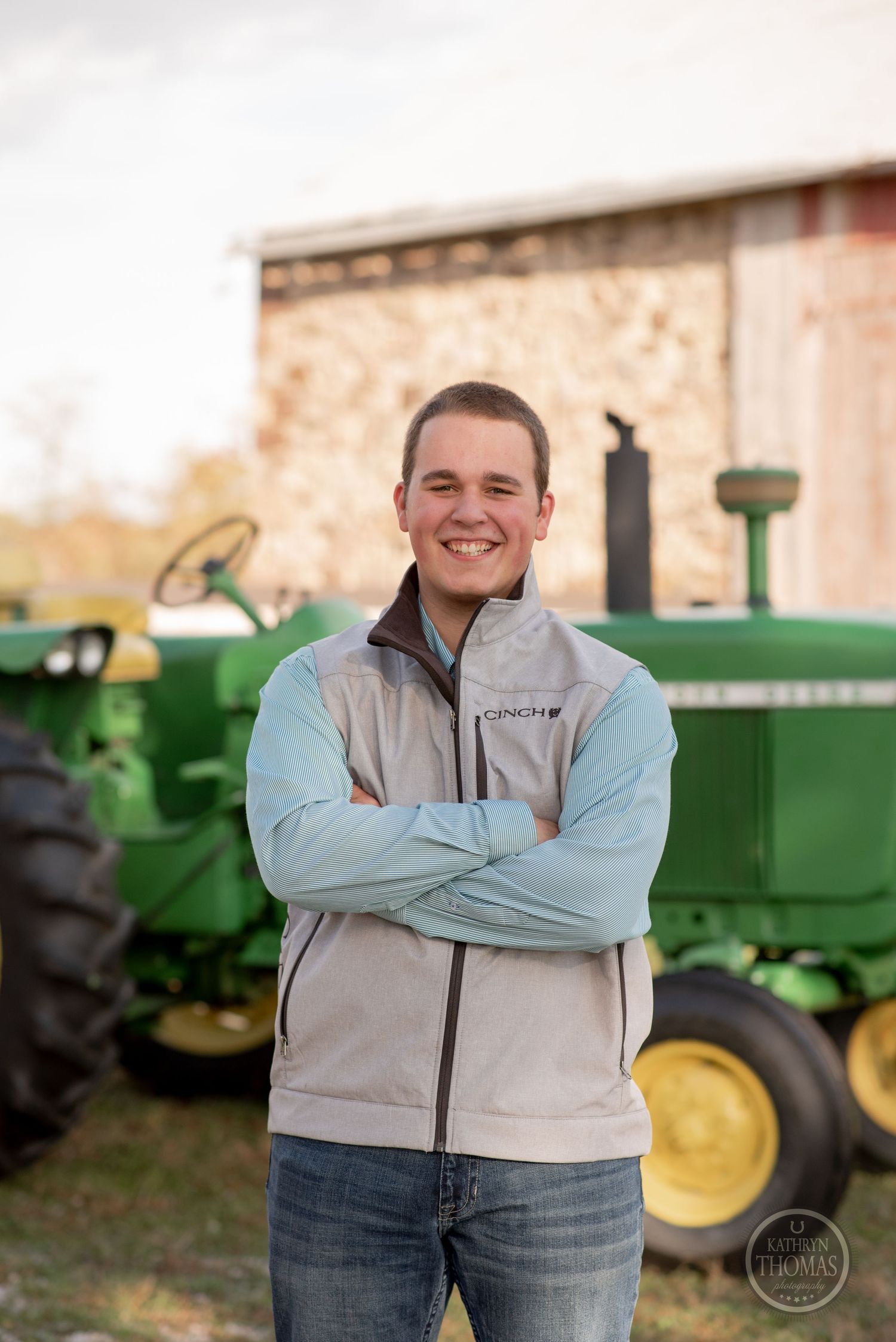 Person wearing light vest and blue shirt poses with crossed arms in front of vintage John Deere tractors.