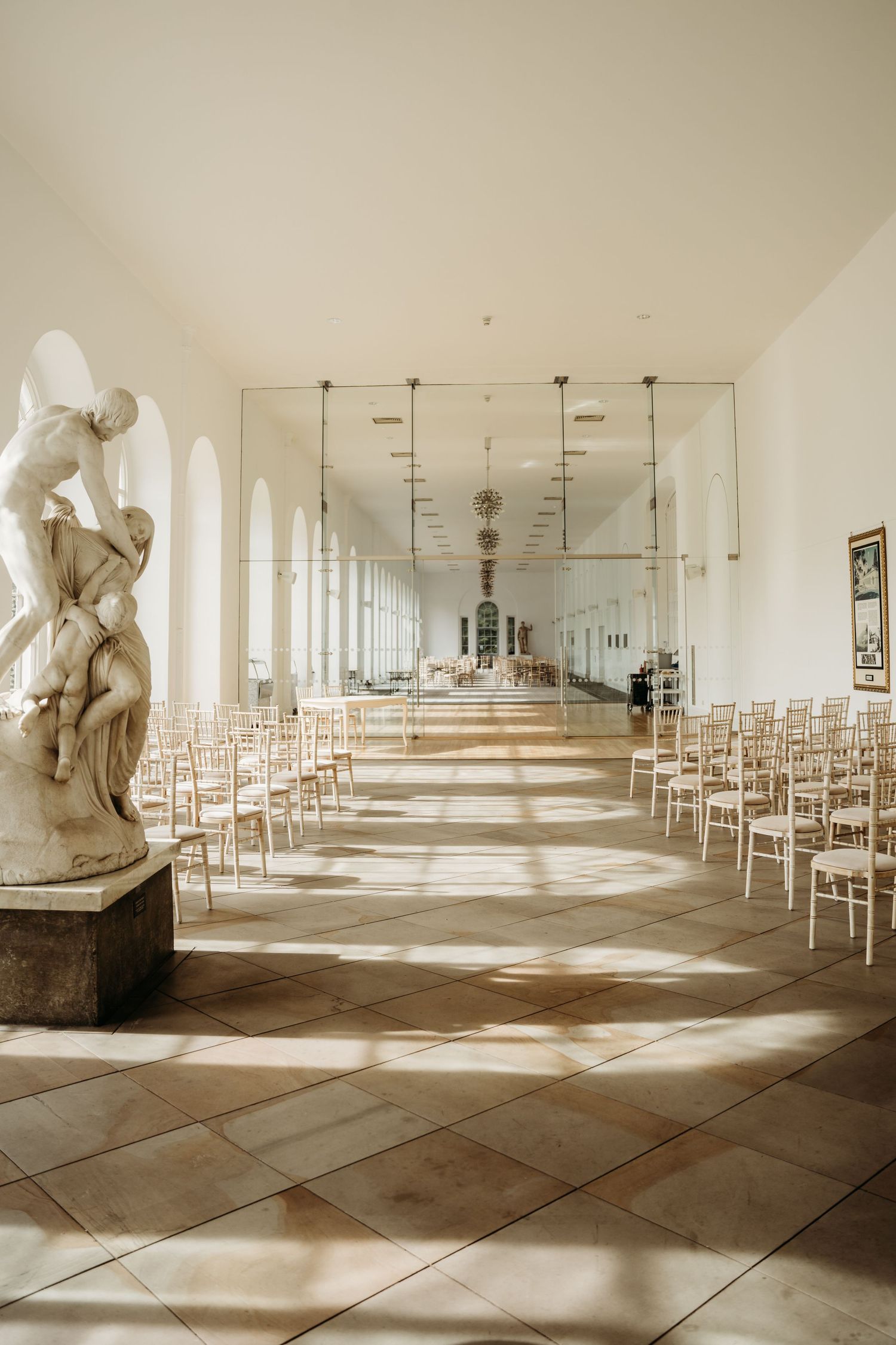 Elegant hallway with classical statue and rows of white chairs illuminated by natural light.